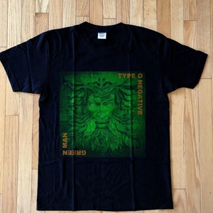 以下が含まれることがあります： 緑と金の葉のついた顔のグラフィックがプリントされた黒いTシャツ。グラフィックには「GREEN MAN」と「TYPE O NEGATIVE」の文字が書かれている。