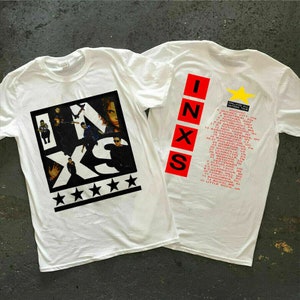 May include: White t-shirt with a black and white graphic of the band INXS. The back of the shirt has a red INXS logo and a list of tour dates for the "Calling All Nations Tour".