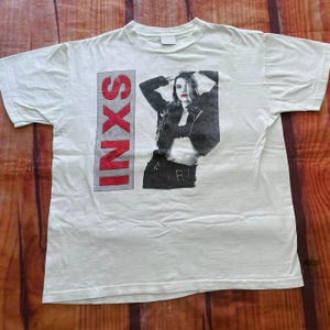 May include: White t-shirt with a red "INXS" logo and a black and white image of a woman wearing a leather jacket and a skirt.