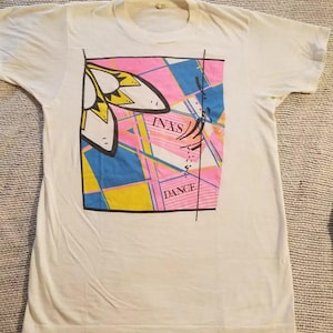 May include: White t-shirt with a colorful graphic design featuring a stylized butterfly, geometric shapes, and the text "INXS DANCE".