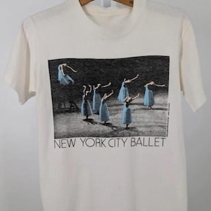 May include: Cream-coloured t-shirt with a monochrome image of ballet dancers in blue dresses. The text "NEW YORK CITY BALLET" is printed below the image. The shirt is hanging on a wooden hanger.