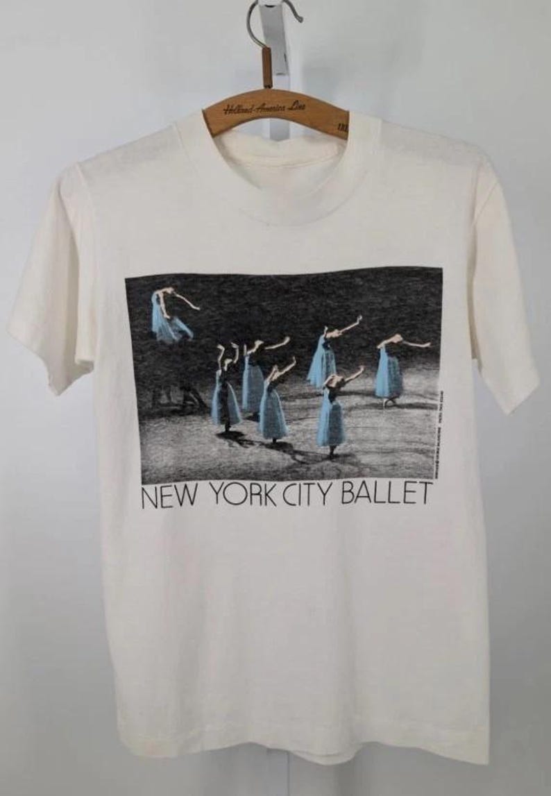 May include: Cream-coloured t-shirt with a monochrome image of ballet dancers in blue dresses. The text "NEW YORK CITY BALLET" is printed below the image. The shirt is hanging on a wooden hanger.