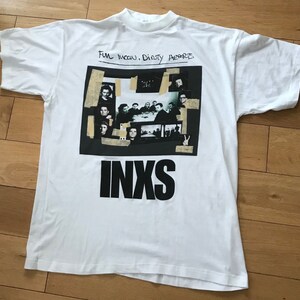 May include: White t-shirt with a black and white photo collage of a band, with the band name "INXS" printed in large black letters below the collage. The text "Full Moon, Dirty Hearts" is handwritten in black marker above the collage.