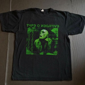 以下が含まれることがあります： 緑色のグラフィックプリントが施された黒いTシャツ。緑色の顔と鎖を持った人物が描かれている。グラフィックの上に「TЯЪЗ O ИЭЯДТИVЭ」というテキストがプリントされている。