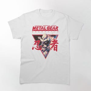 Puede incluir: Camiseta blanca con un gráfico rojo y azul de un personaje del videojuego Metal Gear. El gráfico incluye el texto "Tactical Espionage Action Metal Gear" y caracteres japoneses.