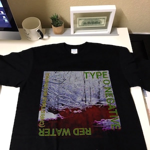 Może przedstawiać: Czarny T-shirt z grafiką przedstawiającą zasneżony las z czerwoną rzeką. Tekst "RED WATER (CHRISTMAS MOURNING)" znajduje się po lewej stronie grafiki, a "TYPE O NEGATIVE" po prawej.