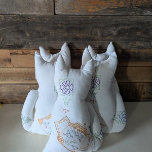 May include: Three white cat-shaped pillows with embroidered floral and teapot designs. The pillows have pointed ears and curved tails. The embroidery features green, purple, orange, and blue thread on the front of the pillows.
