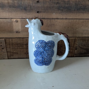 May include: White ceramic pitcher with a rooster design. The pitcher features a blue and white pattern on the front and a brown handle. The rooster's head forms the spout. The pitcher is on a white surface with a wooden background.