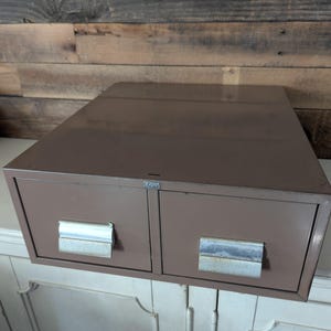 May include: A brown metal file cabinet with two drawers and silver handles. The cabinet is rectangular and sits on a white surface. The background is a wooden wall.