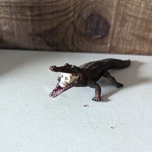 May include: A brown alligator toy with a white skull in its mouth. The alligator has green eyes and is posed with its mouth open.