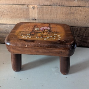 May include: A small, vintage wooden step stool with a rectangular top and four short, rounded legs. The top features a painted illustration of a figure with a pink hat in a field of flowers. The wood has a warm, brown tone.