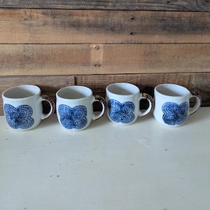 May include: Four white ceramic mugs with blue floral designs and brown handles are arranged in a row. The mugs are on a white surface, with a wooden background. The mugs are likely for coffee or tea.