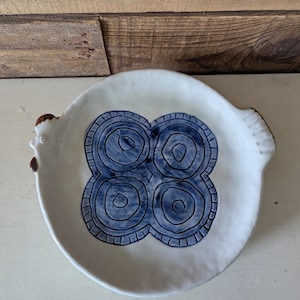 May include: White ceramic serving dish with a blue floral design. The dish has a round shape with two handles and a raised edge. The blue design is composed of four circular patterns. The dish has a rustic, handmade appearance.