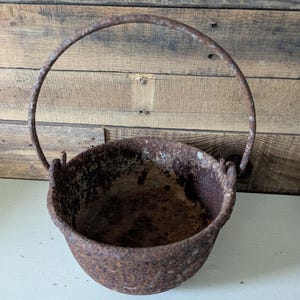 May include: A small, rusty metal cauldron with a rounded body and a sturdy, arched handle. The cauldron's surface shows significant rust, indicating age and exposure. The handle is attached to the sides of the pot, and the interior is dark. The background is a wooden surface.
