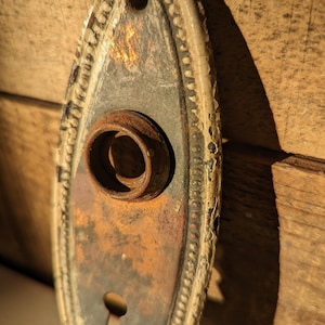 May include: An oval-shaped, antique door knob escutcheon with a keyhole and a round knob hole. The escutcheon is made of metal and has a white painted border with a decorative pattern. The metal is rusted and shows signs of age.