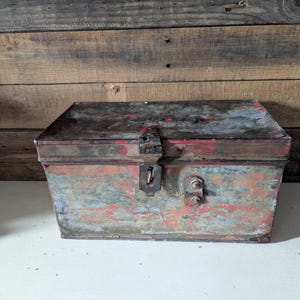 May include: A rectangular metal trunk with a hinged lid, painted in faded red, blue, and gray hues. The trunk features a metal latch and two bolt-like fixtures on the front. The vintage chest sits on a white surface against a wooden background.
