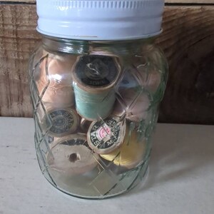 May include: A glass jar with a white lid filled with spools of thread in various colors, including beige, green, yellow, and pink. The jar has a diamond pattern on the sides.