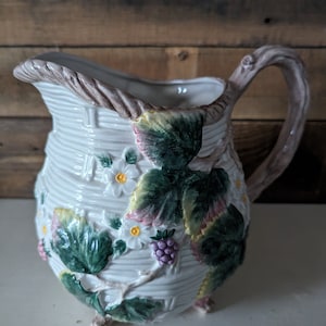May include: White ceramic pitcher with a brown handle and a raised design of green leaves, white flowers, and pink berries.