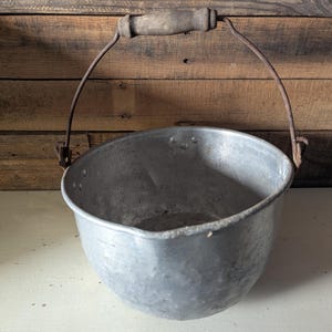 May include: A vintage metal bucket with a curved body and a wooden handle. The bucket has a rustic, aged appearance, with visible wear and tear. The handle is attached by metal brackets. The background is a wooden wall.