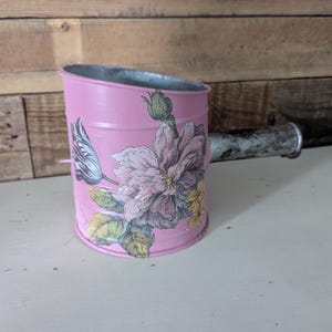 May include: A pink metal watering can with a floral design. The can has a long handle and a spout. The floral design features pink, yellow, and green flowers.