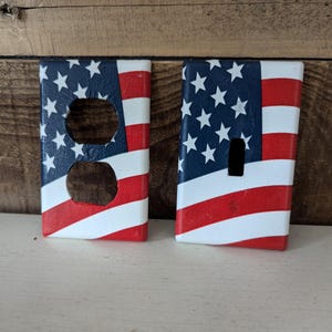 May include: Two decorative wall plates featuring an American flag design. One plate has a double outlet, while the other has a single light switch.