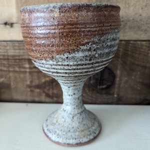 May include: A handmade ceramic goblet with a wide, textured bowl and a slender stem. The goblet features a speckled, off-white base and stem, transitioning to a brown and white striped pattern at the bowl's rim. The goblet is approximately 5 inches tall.
