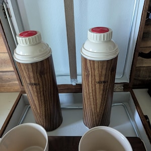 May include: A vintage picnic set featuring two Thermos brand vacuum flasks with woodgrain patterns, cream-colored lids and red stoppers. Two cream-colored cups are also included. The set is inside a brown case with a white interior.