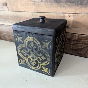 May include: Black wooden box with a gold patterned design and a black knob on the lid.