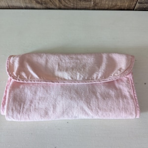 May include: A pale pink fabric baby changing mat with a folded flap and a white seam around the edge.