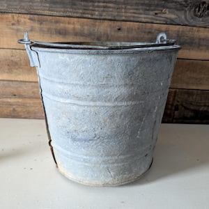 May include: A weathered, galvanized metal bucket with a rounded base and a wire handle. The bucket has a cylindrical shape with vertical ridges and a slightly flared rim. The metal has a dull, gray appearance with signs of age and use.