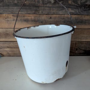 May include: A white enamel bucket with a dark rim and a wire handle. The bucket has a slightly tapered shape and shows signs of age with rust around the base and rim. The background is a wooden wall.