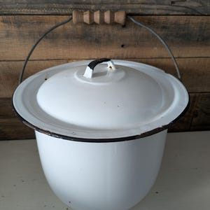 May include: A white enamelware pot with a lid and a wire handle with a wooden grip. The pot has a black rim and a black handle on the lid. The background is a wooden wall.