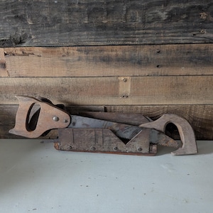 May include: An antique wooden miter box with two hand saws. The saws and box are made of wood and metal, showing signs of age and use. The background is a weathered wooden wall.