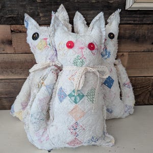 May include: Three white fabric cat toys with patchwork fabric and red button eyes. The cats are sitting on a white surface.