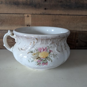May include: White ceramic chamber pot with a floral design in shades of pink, yellow, and green. The pot has a gold rim and ornate detailing.