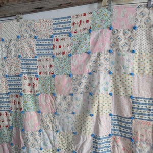 May include: A patchwork quilt with a variety of floral and patterned fabrics in pastel colors. The quilt is adorned with small blue pom-poms at the intersections of the squares. The quilt is hanging from a metal hanger.