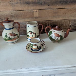 May include: A ceramic tea set featuring a cream-colored base with hand-painted scenes of houses, green trees, and brown roofs. The set includes a lidded pitcher, a creamer, a teapot with a lid, and a single teacup resting on a matching saucer.