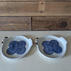 May include: Two white ceramic dishes with blue floral designs. Each dish has a unique shape with small handles and brown accents. The dishes are displayed on a white surface against a wooden background.