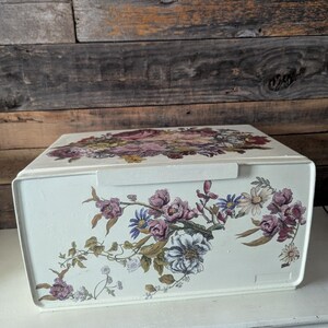 May include: Cream-colored metal bread box with a floral design. The flowers are in shades of pink, purple, white, and yellow. The box has a handle on the top and is sitting on a white surface.