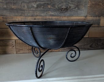 May include: Black metal decorative bowl with a woven design, supported by three scrolled legs. The bowl has an oval shape and sits on a white surface against a wooden plank background. A decorative piece for the home.