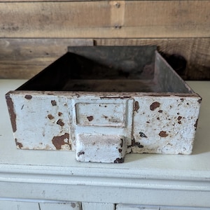 May include: A rectangular metal drawer featuring a distressed white painted finish with visible rust patches throughout. The front panel includes a central rectangular label holder and a protruding handle, revealing a dark, weathered interior.