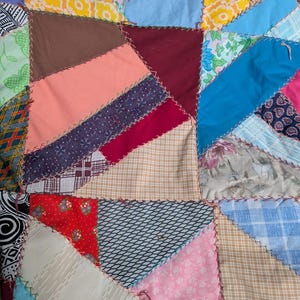 May include: Close-up of a patchwork quilt with various fabric patterns and colors. The quilt features geometric shapes in shades of blue, pink, yellow, brown, and red, stitched together with contrasting thread. A handmade textile.