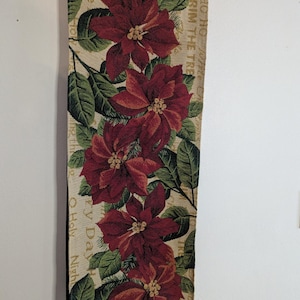 May include: A rectangular Christmas table runner featuring a repeating pattern of large red poinsettia flowers with green leaves on a beige background. The words "HO HO HO" and "TRIM THE TREE" are visible.