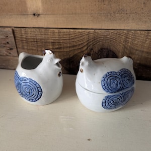 May include: Two ceramic chicken-shaped containers. The white ceramic pieces have blue spiral designs and brown accents. One is open, and the other is closed. The background is a light-colored surface and a wooden wall.