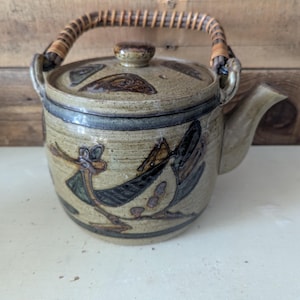 May include: A ceramic teapot with a speckled beige glaze and dark brown hand-painted bird motifs. The teapot features a matching lid, a curved spout, and a woven rattan handle attached to two side loops. The body has horizontal bands.