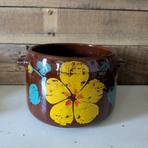 May include: A brown ceramic planter with a yellow flower design. The flower has a red center and green leaves. The planter has two handles on the sides.