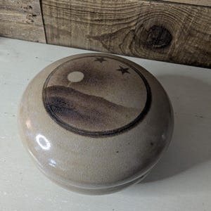 May include: A round, beige ceramic lidded jar with a smooth, glossy finish. The lid features a circular dark brown illustration of a landscape with a sun or moon and two small stars, enclosed within a dark brown circular border line.
