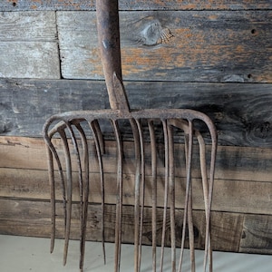 May include: An antique, rusty metal pitchfork with a long handle and multiple tines. The pitchfork is set against a weathered, wooden background, showcasing its aged appearance and rustic charm. The tines are long and slightly curved.