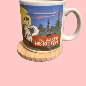 May include: White ceramic mug with a cartoon illustration of a cat in a forest setting. The mug has a red sign that reads "He asked his kitten". The mug is sitting on a wooden coaster.