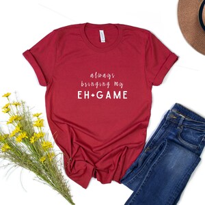May include: Red t-shirt with white text that reads "always bringing my EH*GAME". The shirt is laid flat on a white surface with a pair of blue jeans and a brown hat.